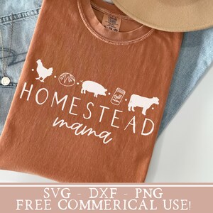 Op de afbeelding: Een bruin T-shirt met de tekst "Homestead mama" in wit. Het ontwerp bevat een kip, een brood, een varken, een pot jam en een koe.