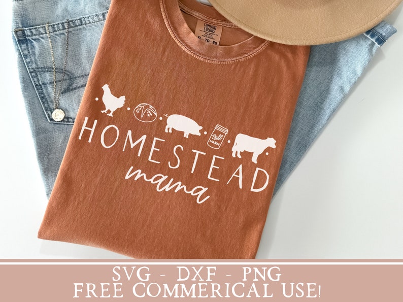 Op de afbeelding: Een bruin T-shirt met de tekst "Homestead mama" in wit. Het ontwerp bevat een kip, een brood, een varken, een pot jam en een koe.