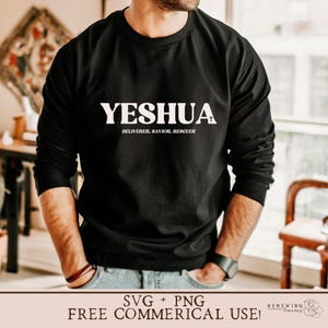 May include: A black long-sleeved shirt with the word "YESHUA" printed in white on the front. The text is followed by a small white cross and the words "DELIVERER, SAVIOR, RESCUER" in smaller white text. The shirt is being worn by a person with their hands in their pockets.