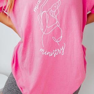 May include: A pink t-shirt with a white outline of a pregnant woman and the text "Motherhood is ministry".
