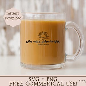 May include: A clear glass mug filled with a light brown liquid, featuring the text "Stay salty. Shine bright." with a sun and wave design. The mug has a handle and is suitable for hot beverages. The text also includes the Bible verse Matthew 5:13-16.