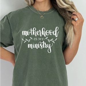 Pu&ograve; includere: Una maglietta verde con il testo "motherhood is my ministry" in lettere bianche con piccoli accenti floreali bianchi.