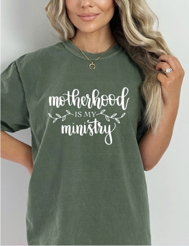 Pu&ograve; includere: Una maglietta verde con il testo "motherhood is my ministry" in lettere bianche con piccoli accenti floreali bianchi.