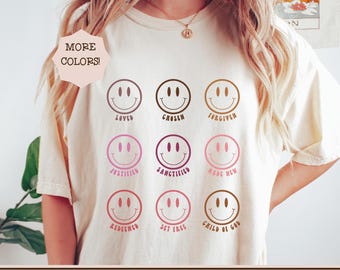 May include: A cream-coloured t-shirt with nine smiley face designs in shades of brown and pink. Each smiley face has a different word underneath, including "Loved", "Chosen", and "Forgiven". The text "MORE COLORS!" is in a pink hexagon.