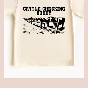 以下が含まれることがあります： 牛がフェンスのある牧場にいて、「Cattle Checking Buddy」という文字が書かれた白いTシャツです。