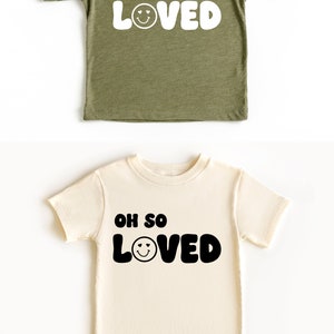 以下が含まれることがあります： 「Oh so loved」という文字と笑顔の顔が付いた幼児用Tシャツ2枚。Tシャツは色が異なり、1枚は緑色、もう1枚はクリーム色です。