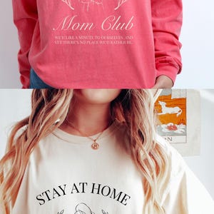 May include: Two images of a white t-shirt with a black graphic design. The graphic design features a woman holding a child with the text "Stay at Home Mom Club" and "We'd like a minute to ourselves, and yet there's no place we'd rather be."