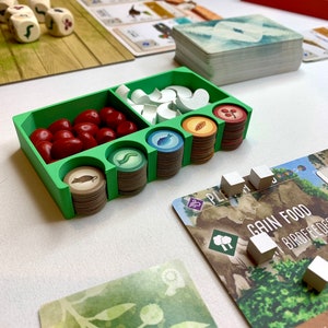 May include: A green plastic organizer with compartments for game pieces. The organizer contains red and white game pieces, as well as stacks of brown, green, and blue tokens. The text "Gain Food" is visible on a brown card.