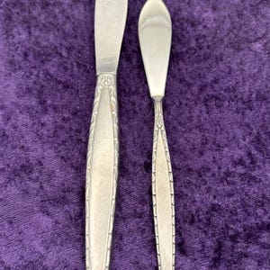 May include: Two silver butter knives with ornate handles. The larger knife has a pointed tip, while the smaller knife has a rounded tip.