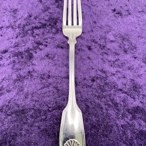 May include: A silver-toned metal dinner fork featuring a four-tined head and a long, slender handle. The handle is decorated with a vertical ribbed pattern and terminates in a raised, embossed shell motif at the base of the utensil.