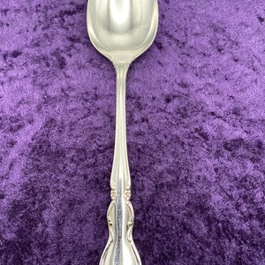 May include: A polished silver-tone metal spoon featuring an elongated oval bowl and a slender handle. The handle is decorated with embossed floral details and curved lines that widen toward the base, creating a symmetrical, ornate appearance.