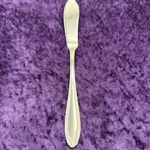 May include: A silver butter knife with a pointed tip and an ornate handle. The knife is on a purple velvet background.