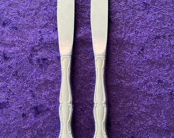 May include: Two silver dinner knives with ornate handles on a purple background.