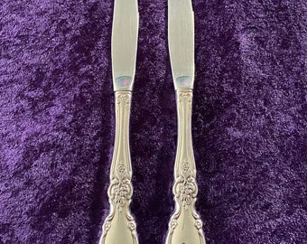 May include: Two silver dinner knives with ornate handles on a purple velvet background.