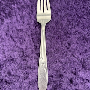 May include: A stainless steel fork with four tines, resting on a purple velvet surface. The handle features a decorative star and curved lines. This utensil is designed for eating and is a common item in tableware.