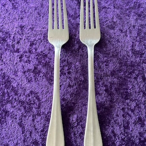 May include: Two silver forks with a polished finish, placed side-by-side on a purple velvet surface.