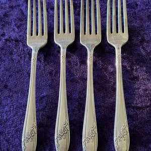 May include: Four silver forks with floral detailing on the handles. The forks have four tines each and are arranged vertically against a deep purple background. The handles have a decorative floral pattern.