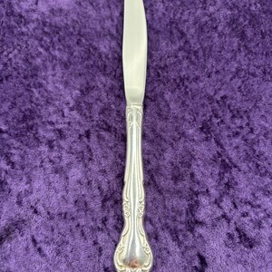 May include: A silver dinner knife with an ornate handle. The knife has a plain blade and a decorative, floral-patterned handle.
