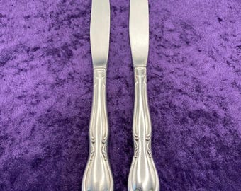 May include: Two silver butter knives with curved blades and ornate handles. The handles feature a detailed, symmetrical design. The knives are displayed against a purple velvet background, creating a contrast of textures and colors. These knives are suitable for formal dining.