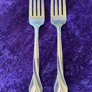 May include: Two silver forks with ornate handles on a purple velvet background.