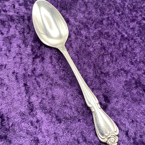 May include: A silver spoon with a rounded bowl and a decorative handle. The handle features a floral design at the base. The spoon is set against a purple velvet background.