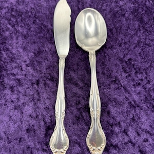 May include: Two silver-plated utensils, a butter knife and a spoon, with ornate handles. The utensils are on a purple velvet background.
