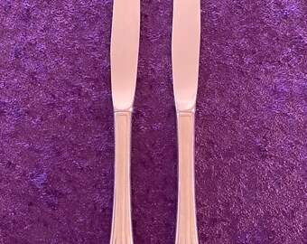 May include: Two silver dinner knives with ornate handles on a purple background.