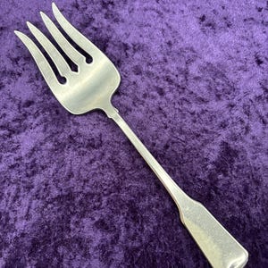 May include: A silver serving fork with four tines and a long, rectangular handle. The fork is made of a shiny metal and is set against a deep purple velvet background. The tines have a slight curve.