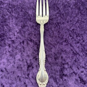 May include: A silver fork with an ornate design on the handle. The fork is on a purple velvet background.