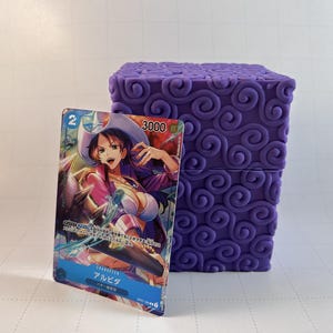 May include: A purple, cube-shaped object with a swirling pattern stands next to a trading card. The card features a character illustration with the number "2" and the number "3000" visible. The card has Japanese text.