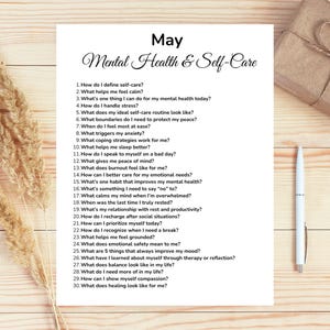 May include: A white sheet of paper with the title "May Mental Health & Self-Care" and a list of 30 self-reflection questions. A pen, a wrapped gift, and dried plants are also on the wooden surface.