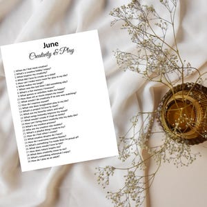 May include: A white paper with the text "June Creativity & Play" and a list of questions. The paper is on a white fabric surface, with a decorative gold-colored bowl and sprigs of white flowers.