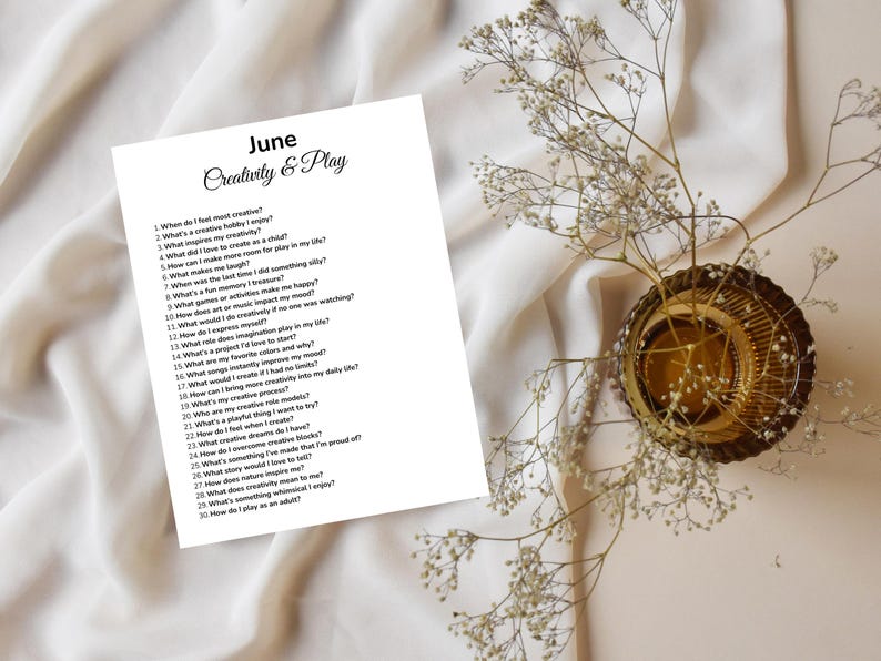 May include: A white paper with the text "June Creativity & Play" and a list of questions. The paper is on a white fabric surface, with a decorative gold-colored bowl and sprigs of white flowers.