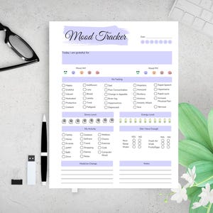 May include: A mood tracker sheet with a lavender header and sections for daily mood, feelings, stress, and energy levels. Includes checkboxes for activities and enough sleep, water, and food. A pen, glasses, and a keyboard are in the background.