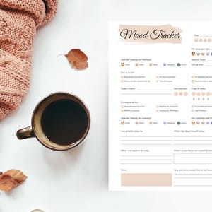 May include: A cozy scene featuring a cup of coffee, a mood tracker, and a soft pink knitted blanket. The mood tracker has sections for daily tasks and self-reflection. Dried leaves add a touch of autumn.