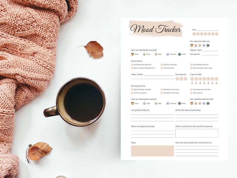May include: A cozy scene featuring a cup of coffee, a mood tracker, and a soft pink knitted blanket. The mood tracker has sections for daily tasks and self-reflection. Dried leaves add a touch of autumn.