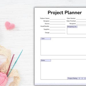 May include: A white Project Planner sheet with the title "Project Planner" at the top. The sheet has sections for pattern details, dates, and notes. Pink yarn balls, crochet hooks, and a pink heart are on the left.