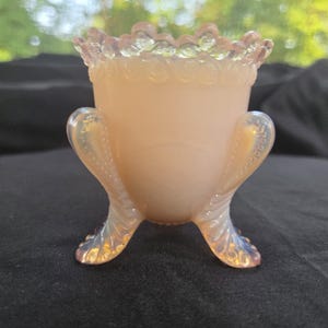 May include: A pale peach-colored glass vessel featuring an opalescent finish, a scalloped rim with a beaded texture, and three curved, ornate feet that extend upward along the sides of the body, creating a distinct tripod base structure.