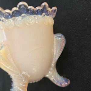 May include: A small, translucent opalescent glass vessel with a creamy white body and a blue-tinted scalloped rim. The piece features three curved, beaded legs and decorative beaded accents along the sides, showcasing a smooth, glossy finish.