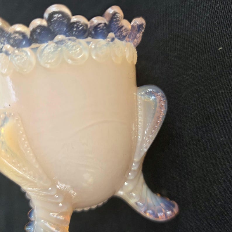 May include: A small, translucent opalescent glass vessel with a creamy white body and a blue-tinted scalloped rim. The piece features three curved, beaded legs and decorative beaded accents along the sides, showcasing a smooth, glossy finish.