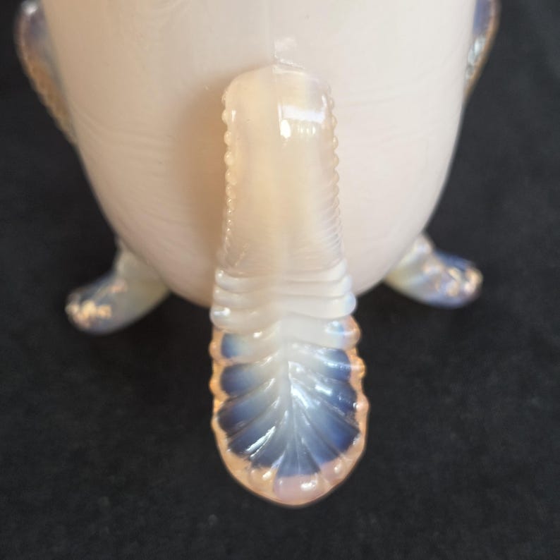 May include: An opalescent glass vessel featuring a milky white body with a subtle iridescent sheen. The piece is supported by three distinct, curved feet that exhibit a translucent blue tint at the tips, mirroring the design of the handle.