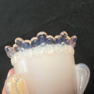 May include: A small, translucent opalescent glass vessel featuring a decorative rim with a repeating floral pattern. The upper edge of the floral design is tinted in a deep blue hue, while the body of the vessel exhibits a milky, iridescent finish.