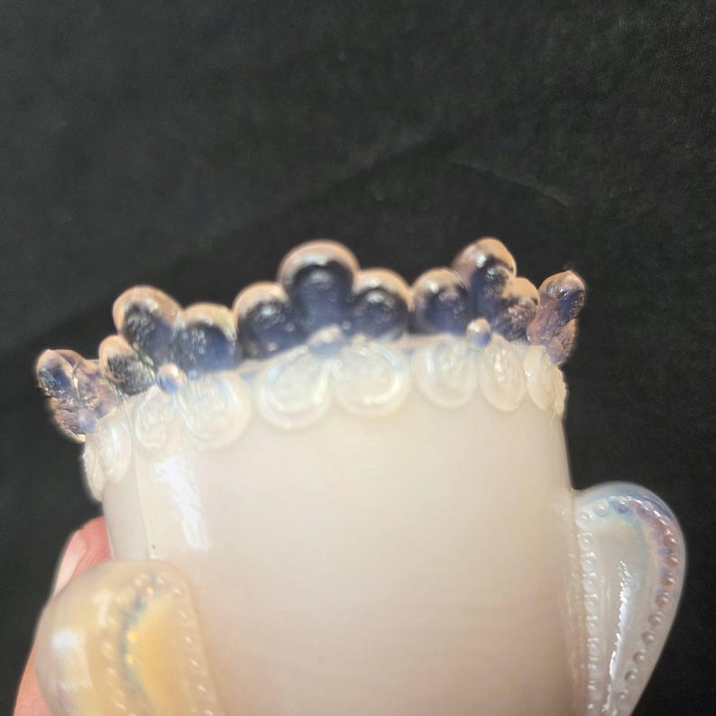 May include: A small, translucent opalescent glass vessel featuring a decorative rim with a repeating floral pattern. The upper edge of the floral design is tinted in a deep blue hue, while the body of the vessel exhibits a milky, iridescent finish.