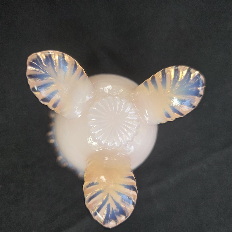 May include: A pale, translucent opalescent glass bowl featuring three distinct feet with a ribbed, leaf-like pattern. The base of the bowl displays a central circular sunburst or starburst design, with the glass exhibiting a soft, milky white hue.