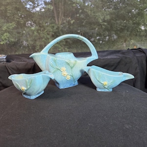 May include: A light blue ceramic tea set consisting of a teapot with a high arched handle and two matching open bowls. Each piece features a raised relief design of green stems and small yellow flowers against a smooth, glossy glazed finish.