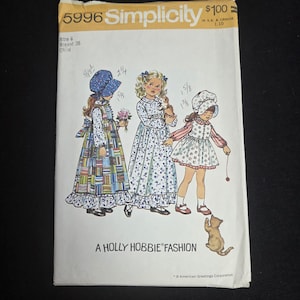 Op de afbeelding: Een Simplicity 5996 naaipatroon envelop voor een kind, maat 6, borstomvang 26, Holly Hobbie mode. De hoes bevat illustraties van drie kinderen in lange jurken en mutsjes, met handgeschreven notities en een kleine illustratie van een kat.