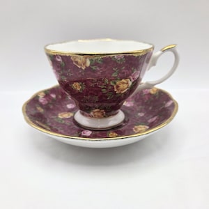 Puede incluir: Un juego de taza de té y platillo de porcelana con un fondo de color burdeos intenso y un patrón repetitivo de rosas amarillas y rosas. El borde tanto de la taza como del platillo está acentuado con un fino ribete dorado y el interior es blanco.