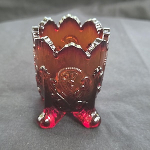 May include: A deep amber-colored glass toothpick holder featuring a scalloped top edge and three small feet. The exterior surface is decorated with a raised, embossed paisley-like pattern that catches the light, creating a translucent effect.