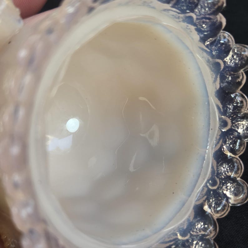 May include: A translucent opalescent glass bowl with a smooth, rounded interior basin and a decorative outer rim featuring a repeating pattern of small, raised circular beads. The glass exhibits a milky, pearlescent sheen with soft blue highlights.