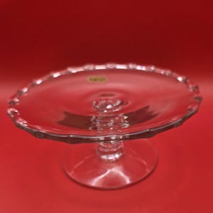 Puede incluir: Un soporte para pasteles de vidrio transparente con un borde festoneado. El soporte tiene una base redonda y un tallo central que sostiene el plato. El fondo es rojo liso.